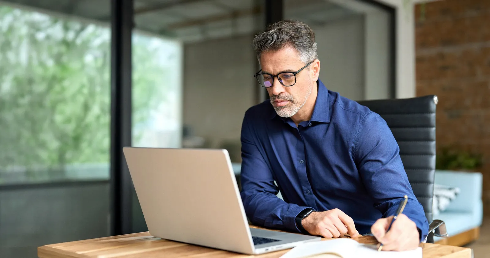 Mann im Hemd arbeitet konzentriert am Laptop im Büro und macht sich Notizen