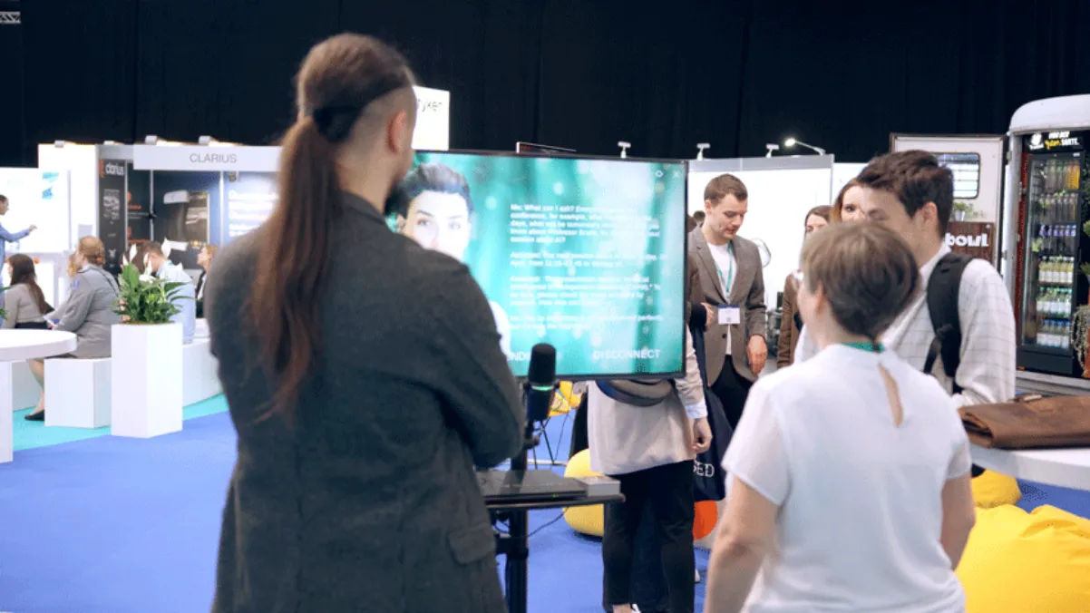 Uczestnicy DCK 2024 w Lipsku stoją przed ekranem z asystentem AI i mówią do mikrofonu